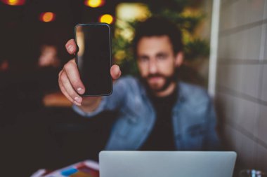 Elinde siyah ekran olan dijital akıllı telefon cihazına odaklan. Kapalı mekanda oturan genç adam. Hippi adam modern cep telefonunu tutuyor ve bunu kameraya internet uygulaması ile gösteriyor.
