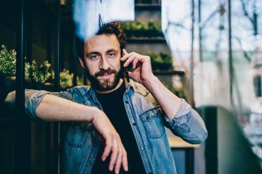 Modern iş yerinde akıllı telefondan iletişim kuran çekici, sıradan giyinmiş genç bir adamın portresi. Sakallı yakışıklı hippi bir öğrenci arkadaşıyla konuşmak için cep telefonundan arıyor.