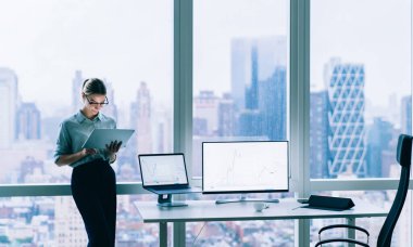 Gündüz vakti büyük ofis penceresinin arkasında bilgisayarlarla duran tablet kullanan zarif, modern kadın.