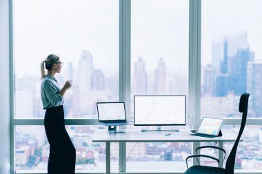 Gözlüklü, zarif, rahatlatıcı bir kadının bir fincan kahve içerken şehir manzarası olan büyük pencereye baktığı yan manzara.