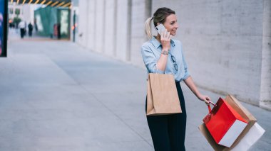 Neşeli, genç bir kadın, siyah cuma günü satışta markalı giysiler aldı, mutlu bir kız sokakta kağıt torbalarla yürürken akıllı telefon cihazıyla iletişim kuruyor, 4G kablosuz internet kullanıyor.