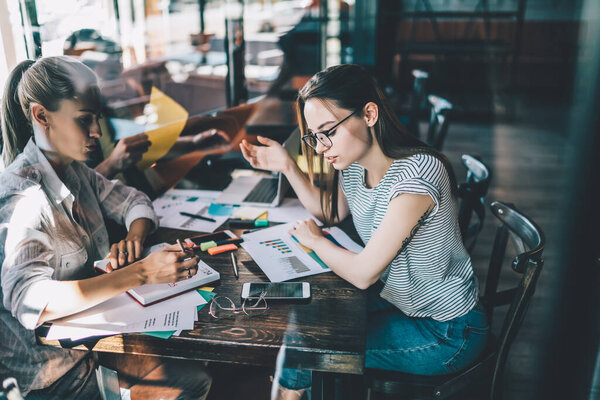 Side view through glass of casual women with notepad and chart sitting at table in cafe and working in team