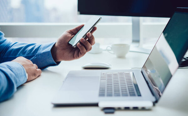 Faceless side view of modern businessman watching mobile phone while sitting at table with laptop and computer against window in office