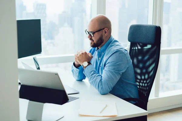 专注的秃头男人 戴着眼镜和蓝色衬衫 手牵手坐在现代办公室的电脑桌前 看着笔记本电脑 — 图库照片