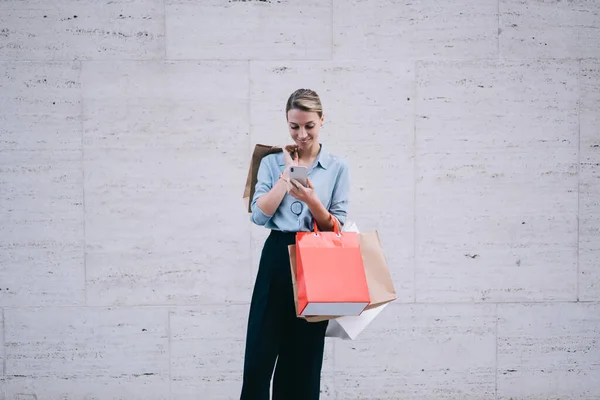 Happy female using mobile phone for sending text messages in social network sharing info about sales during Black Friday, millennial woman holding smartphone and paper bags near advertising area