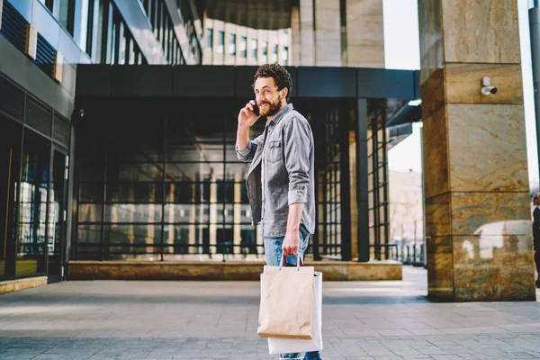 Handsome caucasian male buyer satisfied with delivery holding shopping bags with copy space for advertising text or label, smiling hipster guy enjoying purchases calling via cellular in store