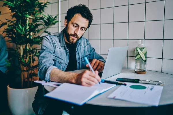 Pensiv Skäggig Ung Man Studerar Bärbar Dator Och Skriva Ner — Stockfoto