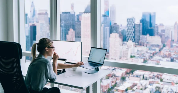 ラップトップを使用しながら大きな窓のある現代的なオフィスのテーブルに座って眼鏡でスタイリッシュなブロンドの女性の側面図 — ストック写真