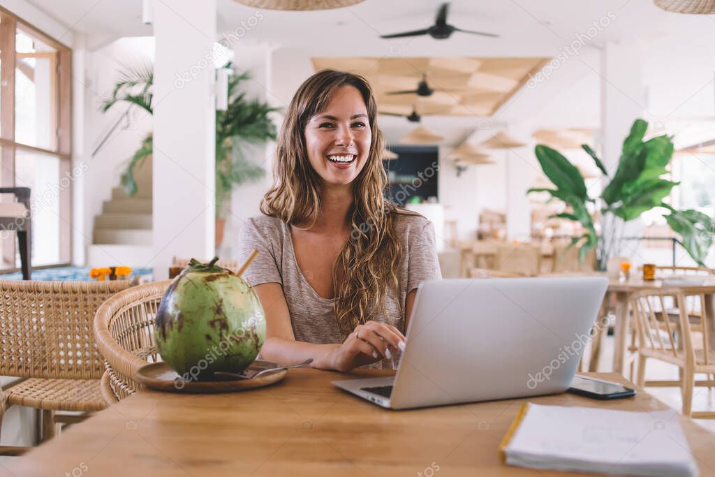 Portrait of excited hipster girl in casual wear laughing and enjoying leisure day in cafeteria using laptop computer for online blogging , positive Caucasian digital nomad at table with tropical fruit