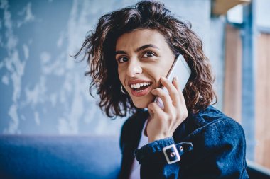 Şık, kıvırcık saçlı, kısa saçlı, modern akıllı telefon cihazıyla gülümseyen ve konuşan bir kadın. Sıradan giyimli, çekici, hippi bir kız dijital cep telefonundan arıyor ve başka tarafa bakıyor.