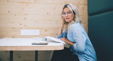 Moda gözlüklü, zeki, beyaz tenli bir kızın, kapalı alanlarda çalışmak için yararlı edebiyat kullanarak üniversite ödevi için bilgi araştırması yaparken kameraya bakması.