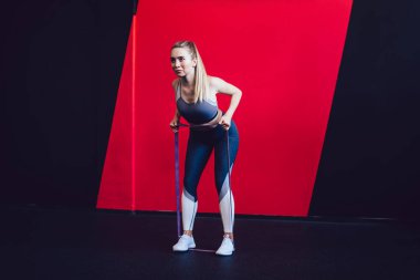 Çekici beyaz kadın sporcu direniş bandıyla egzersiz yapıyor. Önüne bakıyor ve vücut kaslarına konsantre oluyor. Modern spor salonunda mükemmel şekil verme eğitimi olan genç güçlü kadın.