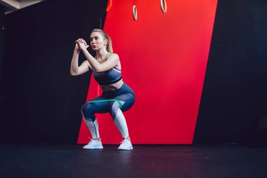 Attractive Caucasian fit girl training legs and ass muscles in sport hall squatting with elastic band, beautiful woman doing crouch during workout using elastic expander for exercising indoors