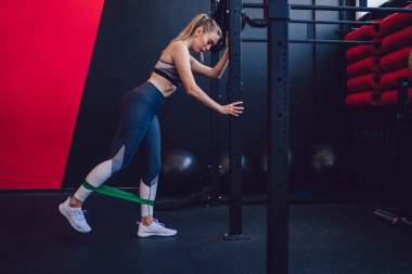 Beyaz kadın antrenör sportif kısa etek ve spor tozluklarıyla fitness lastik bantlarla hamleler yapıyor, çekici bayan atlet pilates stüdyosunda elastik genişletici ile bacaklarını açıyor.
