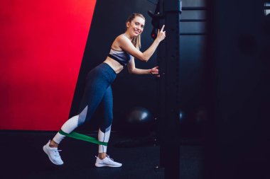 Eşofmanlı neşeli beyaz kadın sporcunun tam boy portresi kameraya gülümserken bacak kaslarını elastik genişletici ile ısıtıyor, mutlu bir kadın spor salonunda yeşil bant kullanıyor.