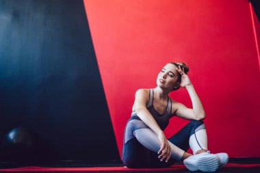 Eşofmanlı düşünceli bir bayan sporcu spor salonundaki kardiyo eğitiminden sonra dinleniyor pilates halısında oturuyor ve arka planda promosyon duvarı var, vücut geliştiricinin bakışlarını kaçırdığını düşünüyor ve düşünüyor.
