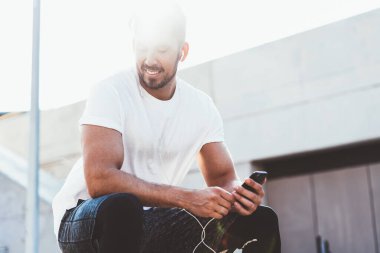 Başarılı erkek koşucunun gülümseyip iyi hissetmesinin altında kent ortamında antrenman yapan mutlu sporcu açık havada oturup kulaklıkla pozitif müzik listesi dinliyor.