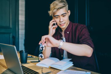 Sarışın, pozitif Çinli genç adam, bilgisayar başında çalışırken akıllı telefondan arama yaparken akıllı saati kontrol ediyor. Japon hippi öğrenci cep saatine bakıyor ve cep telefonuyla konuşuyor.