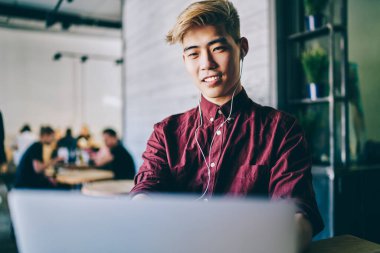 Neşeli Asyalı öğrenci kulaklıklarıyla modern bilgisayarda komik dersler seyrediyor. Modern mekanda oturan Çinli genç adam dijital bilgisayarda kablosuz internet kullanarak serbest çalışıyor.