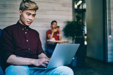 Neşeli Çinli hippi öğrenci modern dizüstü bilgisayarda ders çalışması için günlük giyim metni yazıyor ve birlikte çalışma alanındaki kablosuz internet bağlantısını kullanarak internet sitelerinde arama yapıyor.