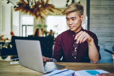 Gülümseyen Japon hipster laptop bilgisayarına kablosuz internet kullanarak program yükleyen geliştirici. Millennial genç adam netbook 'ta internet üzerinden özel ders seyrediyor. Modern uzayda başlangıç üzerine çalışıyor.