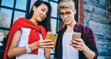 Neşeli hipster Asyalı öğrenciler ellerinde modern akıllı telefonlar ve çevrimiçi video izliyorlar şehir ortamında dikiliyorlar. Pozitif Japon arkadaşlar hücresel ağlarda tasarım profillerini tartışıyorlar.