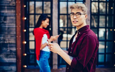 Gözlüklü Çinli hippi, akıllı telefonuyla mesajlaşırken başka tarafa bakıyor ve cep telefonuyla arka planda bulanık duran arkadaşıyla iletişim kurmak için 4G interneti kullanıyor.