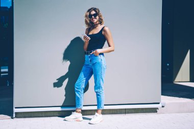 Akıllı telefonlu neşeli yeniyetme kızın uzun bir portresi. Şehir merkezinde kameraya bakıyor. Güneşli yaz günlerinde cep telefonuyla internette gezen güneş gözlüklü mutlu bir kadın.