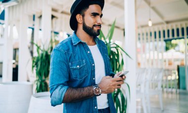 Moda blogunda yayınlanması için elinde cep telefonu cihazıyla gezen modaya uygun giyinmiş İspanyol hippinin yan görüntüsü. Elinde dijital teknoloji olan şık şapkalı olumlu bir adam.