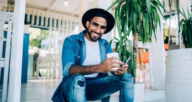 Havalı kıyafetler giymiş mutlu bir adamın portresi. Elinde yemek kupası ve akıllı telefon aleti ile dışarıda oturuyor. Boş zamanlarında kameraya gülümsüyor.