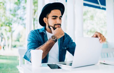 Şık şapkalı düşünceli genç adam internet sitesinden ilginç webinar izliyor sokak kafeteryasında iyi internet bağlantısı olan programcılar için, hipster adam video çağrısı alıyor.