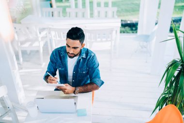 Çevrimiçi internet sitelerindeki ucuz biletleri araştırmak için dizüstü bilgisayarla kafeteryada oturan organizasyon yazarları hakkında bilgi içeren bir ders kitabı tutan deneyimli genç turist.