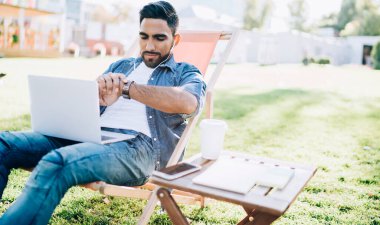 Dizüstü bilgisayar teknolojisiyle uzaktan yakından ilgilenirken takılabilir kol saatini kontrol eden yetenekli erkek serbest yazar, arkadaşı beklerken elektronik kulaklıkla müzik dinleyen genç Türk adam