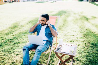 Elektronik kulaklıklar ve dizüstü bilgisayarla müzik dinleyen yakışıklı bir adam, üniversite kampüsünde öğrenim görürken ses kitabına odaklanmış genç bir adam.