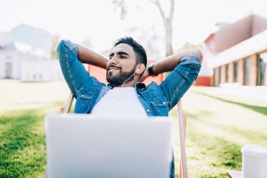 Neşeli erkek meloman boş vakitlerin tadını çıkarıyor ve giyilebilir elektronik kulaklıklarla şehir parkında dizüstü bilgisayar, zevk ve dinlenme konseptiyle dinlenen pozitif müzik listesi dinliyor.