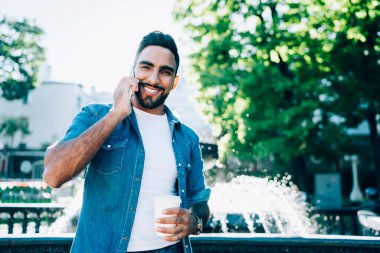 Kahve fincanı tutan ve kameraya gülümseyen şık kot gömlekli neşeli bir erkeğin yarım boy portresi. Bu arada arkadaşlarıyla iletişim kurmak için gezinen interneti kullanıyor.