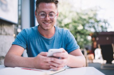 Beyaz tenli pozitif adam internet sitesindeki gençlik yaşam tarzı hakkındaki yorumları kontrol ediyor. Gözlüklü neşeli adam cep telefonuyla mesaj gönderiyor. 4G kablosuz internet bağlantısı var.