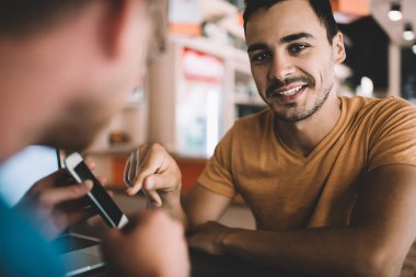 Modern cep telefonu cihazını elinde tutan ve web sitesinde ilginç bilgiler gösteren hipster adamın arka planda görüntüsü. Pozitif erkek arkadaş kameraya bakıp gülümsüyor. İletişim kavramı.
