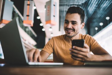 Elinde cep telefonu cihazıyla medya dosyalarını paylaşmak için dizüstü bilgisayardaki bluetooth 'a bağlanan neşeli erkek blogcu, arkadaşının gülümsemesini kontrol eden olumlu hipster adam.