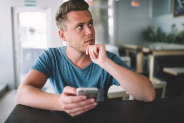 Kafeterya masasında elinde modern cep telefonu cihazıyla oturan yakışıklı hippi sosyal ağ için yeni yayınlar yaratmak için düşünceli bakışlar atıyor.