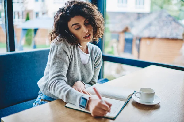 コーヒーを飲みながらテーブルに座っているスマートフォンのクライアントと通信しながら ノートに情報を書く魅力的な若い女性 — ストック写真