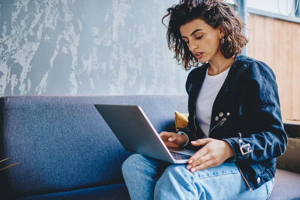 Pensive young woman with short haircut typing text on laptop in search internet app sitting on couch in modern space.Stylish casual dressed hipster girl doing shopping online on web store on netbook