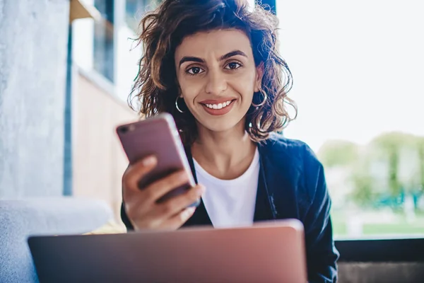 吸引人的高加索年轻女性手持智能手机对着相机微笑 同时还能更新笔记本电脑上博客的形象的肖像 — 图库照片