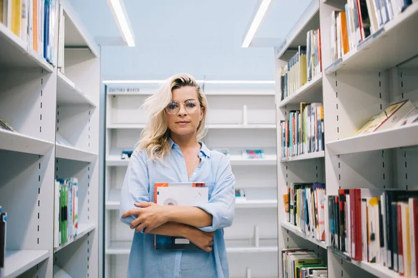 一个有魅力的年轻女子为了读书而在图书馆里呆着看书的画像 一个严肃聪明的女子为了受教育而站在现代书店的书架上 — 图库照片