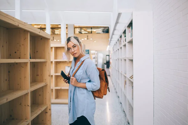 学院里穿着休闲装 手持手机站在书架上的漂亮嬉皮士女孩的半身像 年轻的女学生在大学图书馆里看相机 — 图库照片