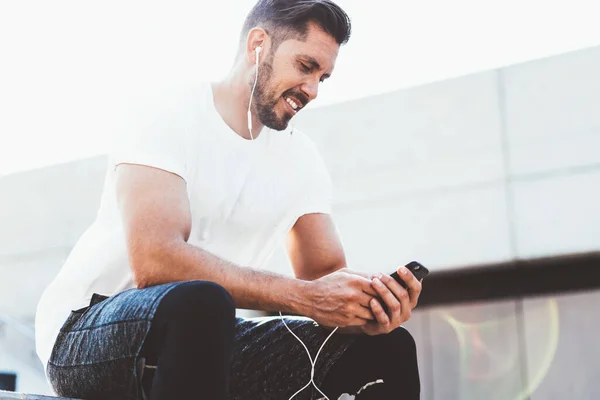 Caucasian male jogger dressed in stylish sportswear reading publication about wright cardio training connected to public internet wireless on modern smartphone gadget,happy man listening audio message