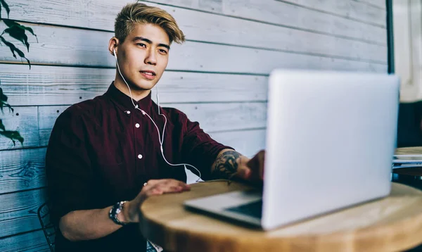 Düşünceli Japon hippi öğrenci kulaklıkla bilgisayarda Webinar 'ı seyrediyor modern birlikte çalışma alanında oturuyor. Konsantre Çinli genç adam internet üzerinden internet üzerinden bilet alıyor.