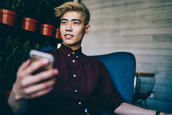Chinese hipster guy dressed in casual wear holding smartphone device in hand while looking away.Handsome japanese young man sending sms message on cellular sitting in coworking space