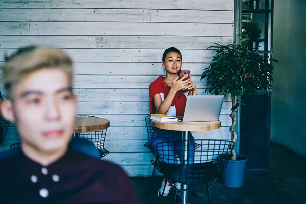 中国の若い女性は スマートフォンのチェックメールを着用し カフェでラップトップコンピュータに座っている友人とオンラインでチャットします — ストック写真