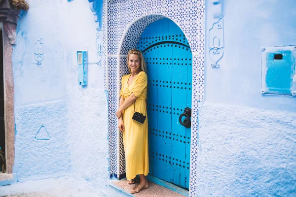 Cheerful hipster girl in casual yellow dress with old fashioned technology on shoulder standing near oriental blue door in Morocco and smiling at camera, concept of travel and discover chaouen culture